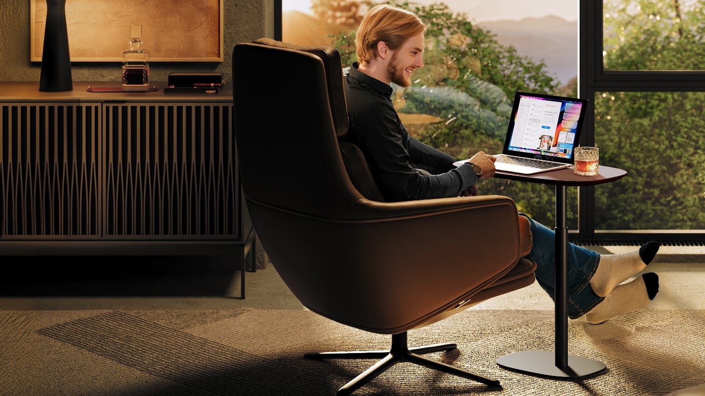 Young man with a beard using a laptop on the BDI Serif 1046 Lift-Top Table while seated in a leather lounge chair, with a beverage nearby.