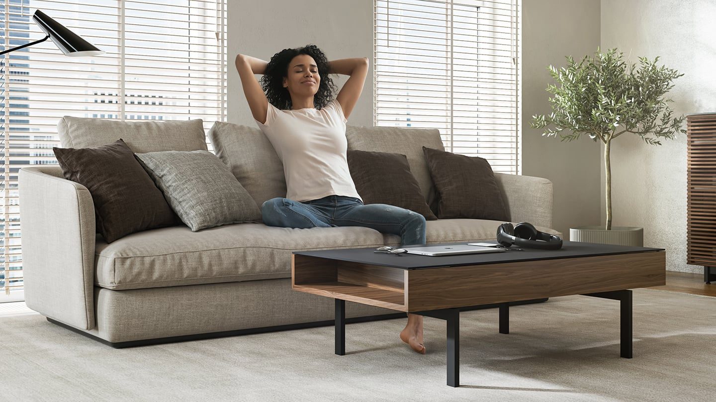 African American woman relaxing on the couch in her new home, sitting in front of a BDI Reveal lift-top coffee table in natural walnut finish.