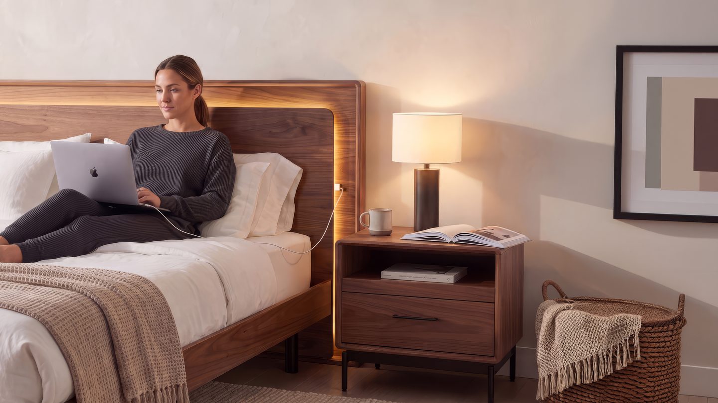 a woman on her computer while on the bed the computer is charged into the headboard